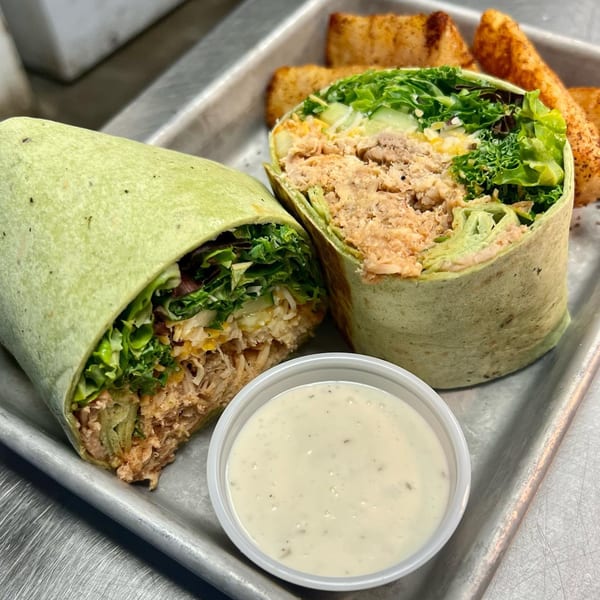 Two halves of a green tortilla wrap filled with chicken and salad, served with a side of ranch and potato wedges.